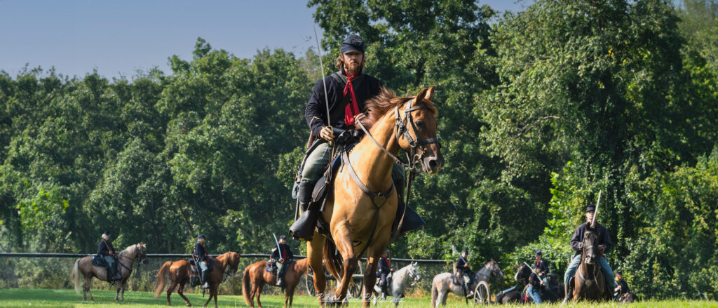 "A Union cavalry soldier sits tall on horseback, leading a line of fellow cavalry soldiers during the American Civil War. Dressed in a blue uniform and holding the reins confidently, the soldier glances ahead, ready for action. Behind him, a formation of cavalry soldiers, similarly mounted and armed, stretches into the distance. Their horses are poised, and the soldiers' sabers or rifles gleam in the light, creating an impressive and disciplined display of military strength."